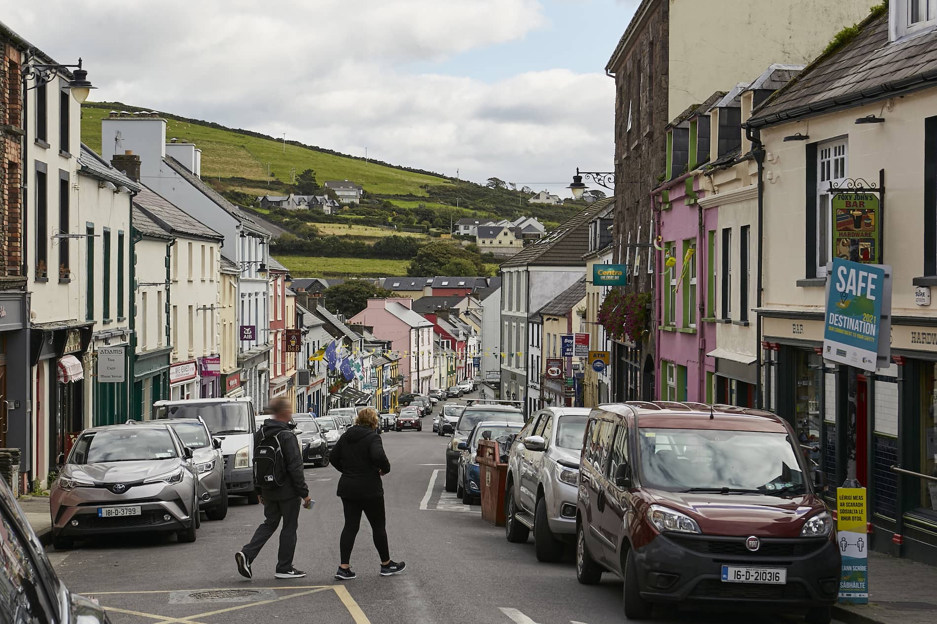 Busy main street in Dingle, County Kerry, with cars parked and people walking past colourful buildings.