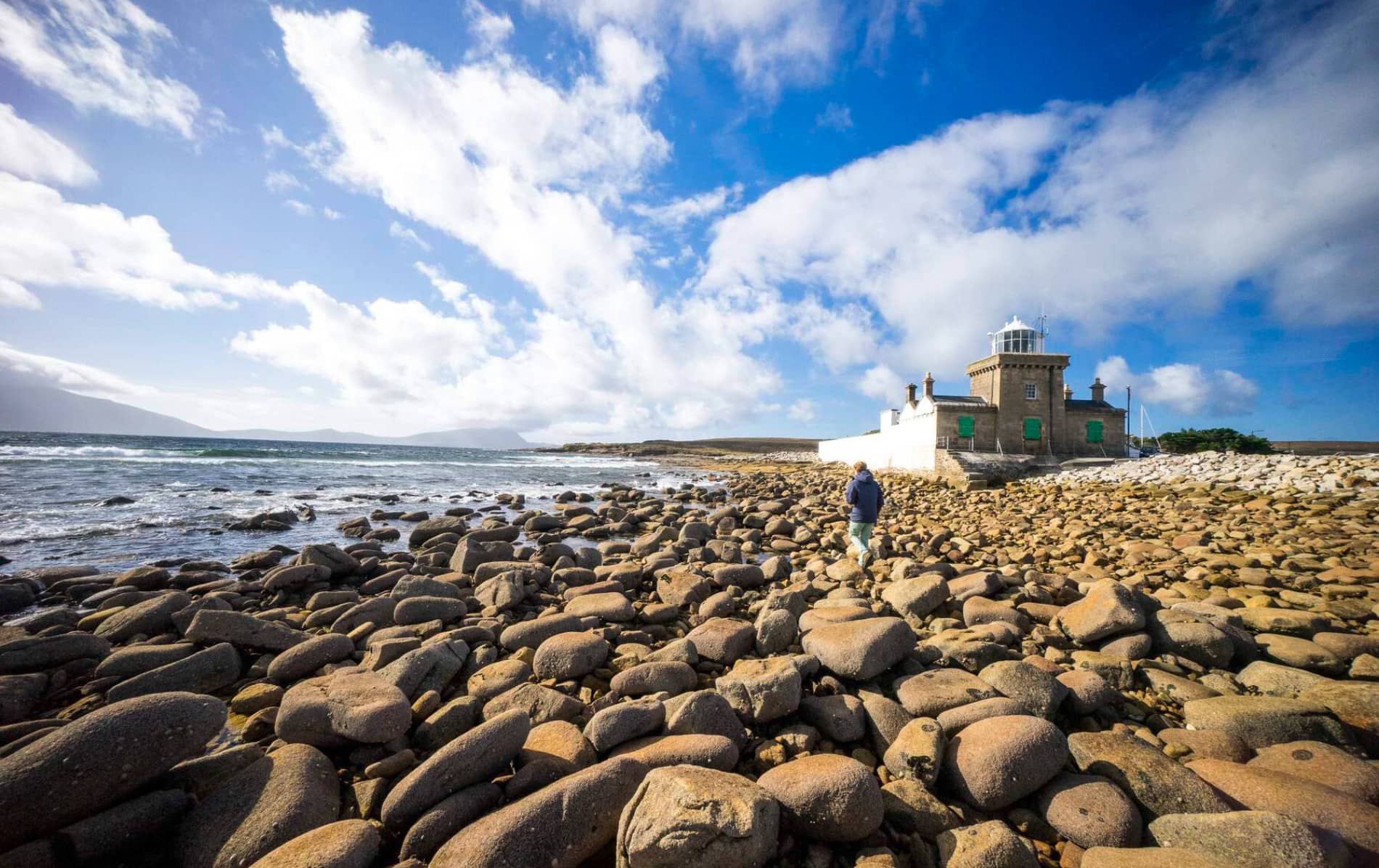 Person walking on boulder-covered shoreline near Blacksod Lighthouse on the Atlantic coast of County Mayo.