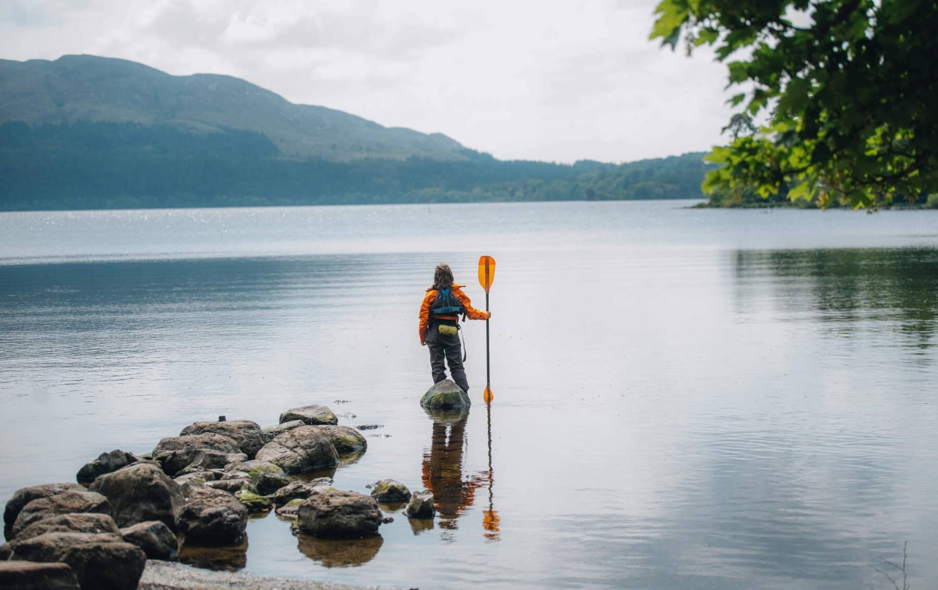 Person in orange jacket holding a paddle while standing on lakeshore rocks in Sligo with mountains in the distance.
