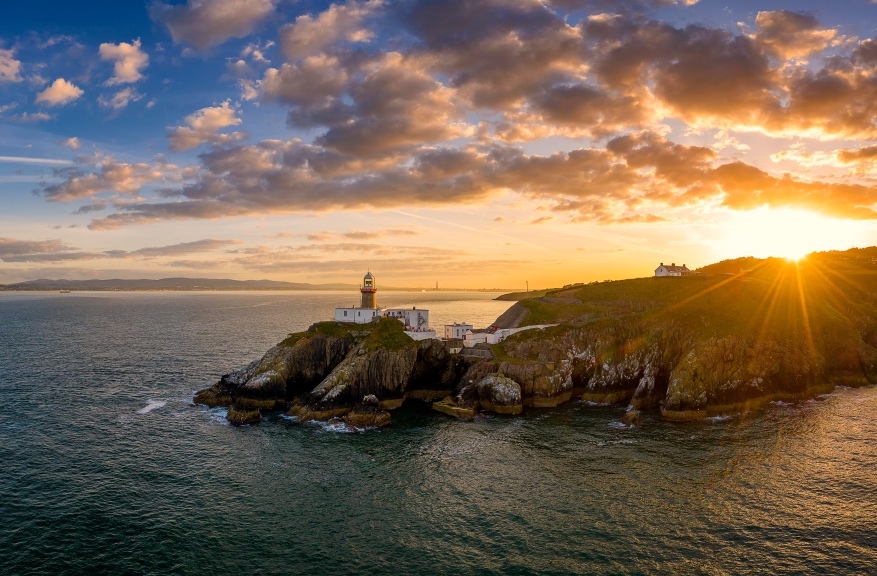 Golden winter sunset over Baily Lighthouse, Howth, Dublin, with sun rays reflecting on calm sea.