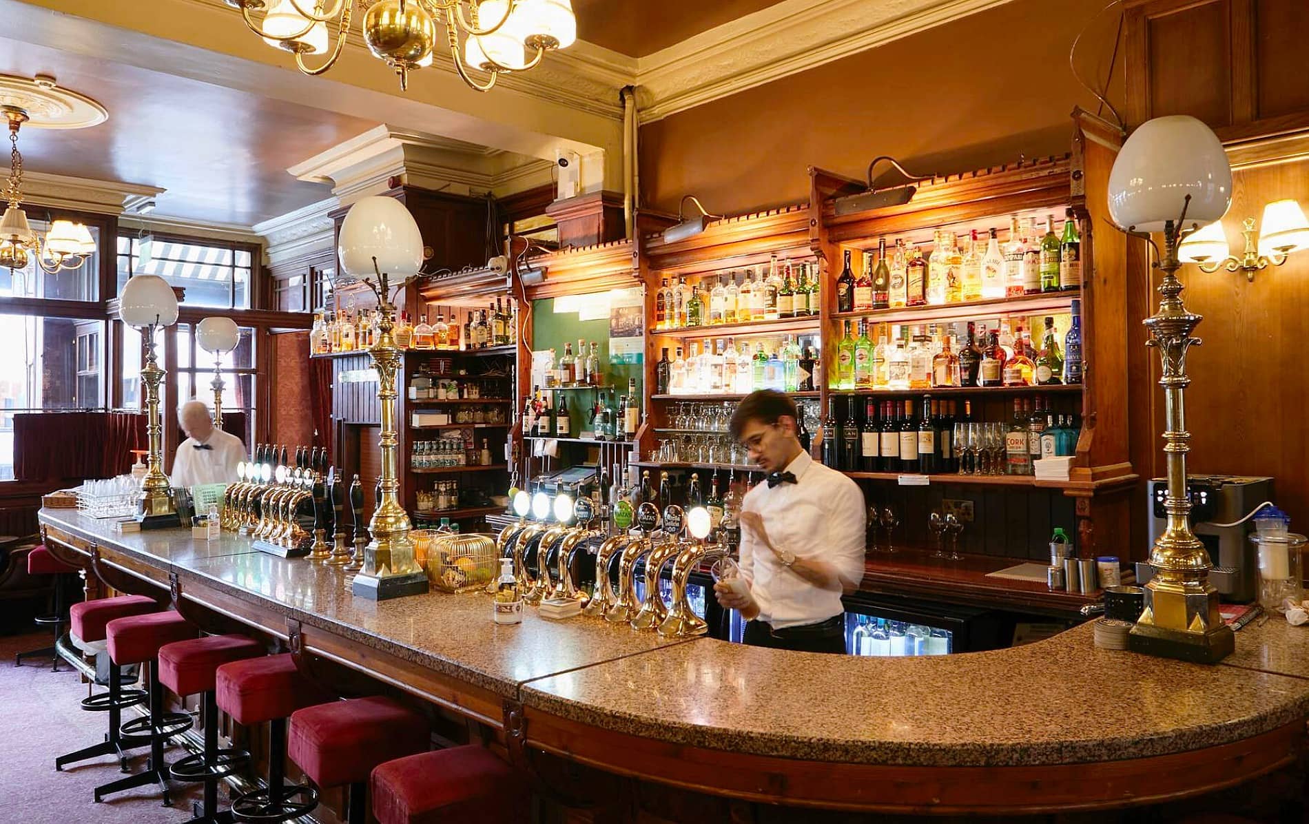 Bartender at Neary’s pub in Dublin prepares drinks behind a polished bar with brass taps and red velvet stools.