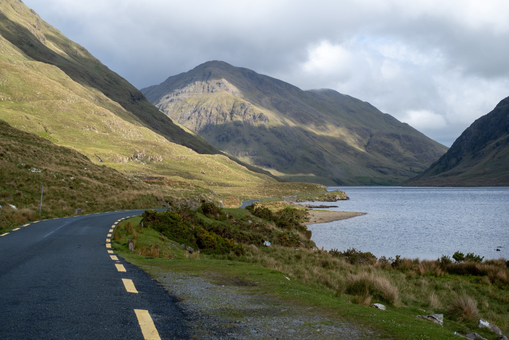 Quiet road winds through the majestic, green slopes of Doolough Valley beside a peaceful lake under cloudy skies.