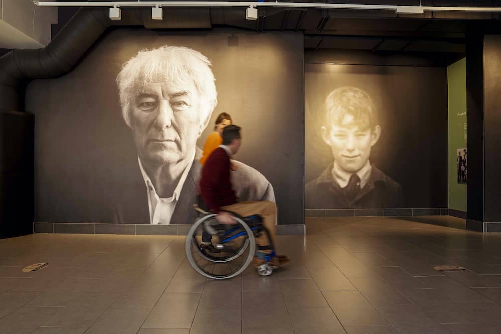 Person in a wheelchair passing portraits of poet Seamus Heaney as adult and child at Seamus Heaney HomePlace, Bellaghy.