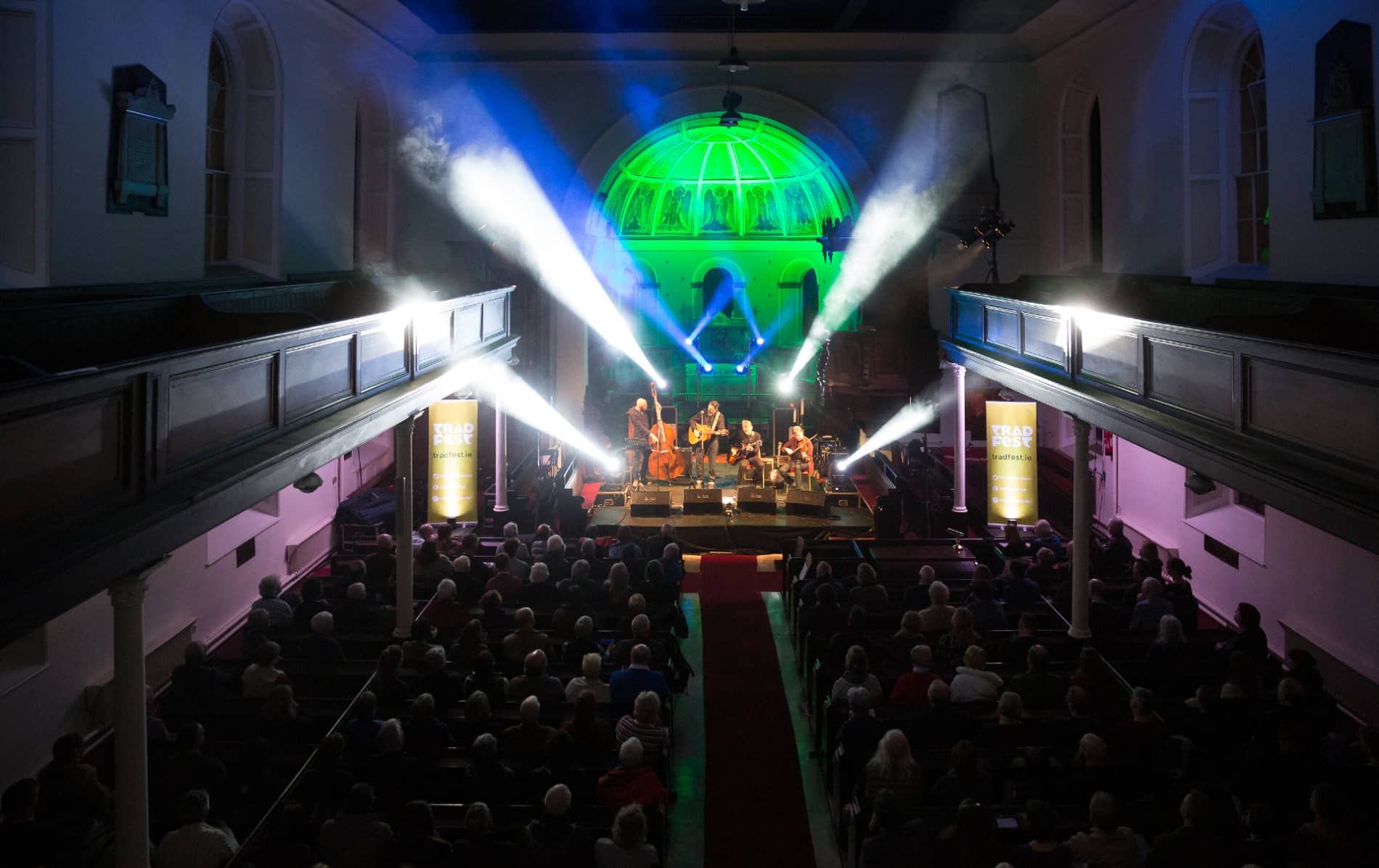 TradFest concert underway in Dublin’s Pepper Canister Church, with a full seated audience and stage lit by vivid beams of light.