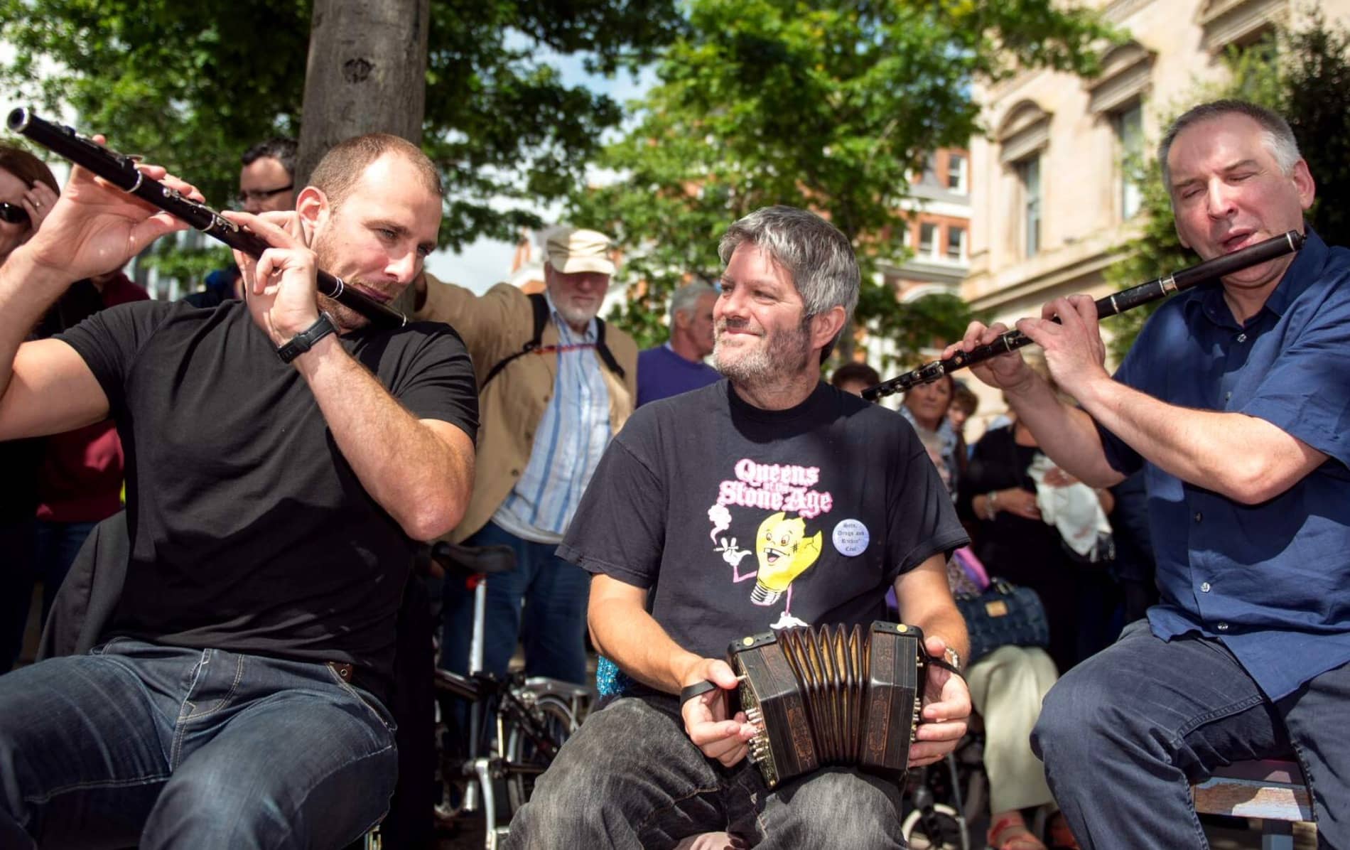 Three musicians play flute and concertina at Fleadh Cheoil, Derry~Londonderry, with a crowd behind them.