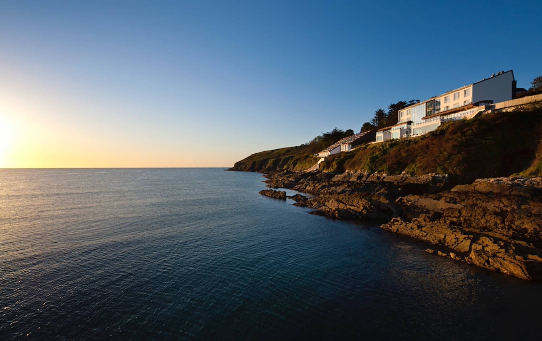 Cliff House Hotel overlooking Ardmore Bay, Waterford, lit by golden sun with rocky coast and calm sea below.