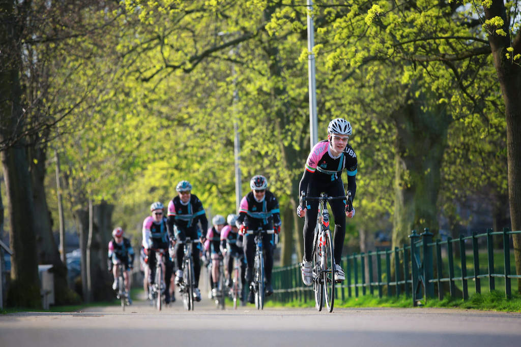 Competitive cyclists in matching kit ride through a tree-lined road in Armagh during a springtime cycling event.