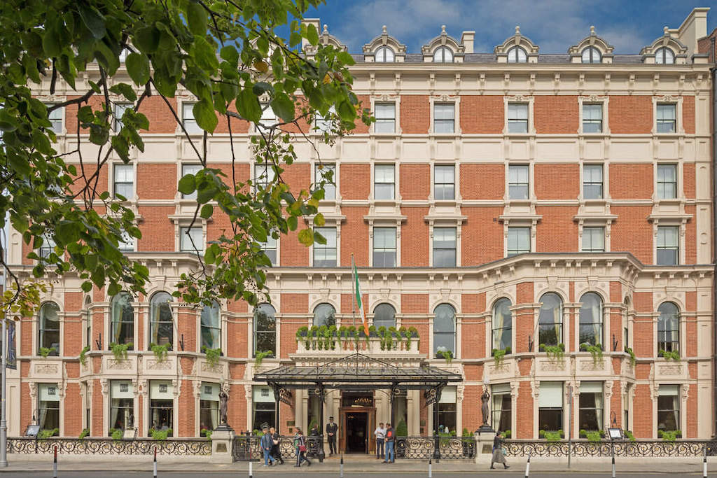 Grand facade of The Shelbourne Hotel on St Stephen’s Green, Dublin, with Irish flag and passers-by at entrance.