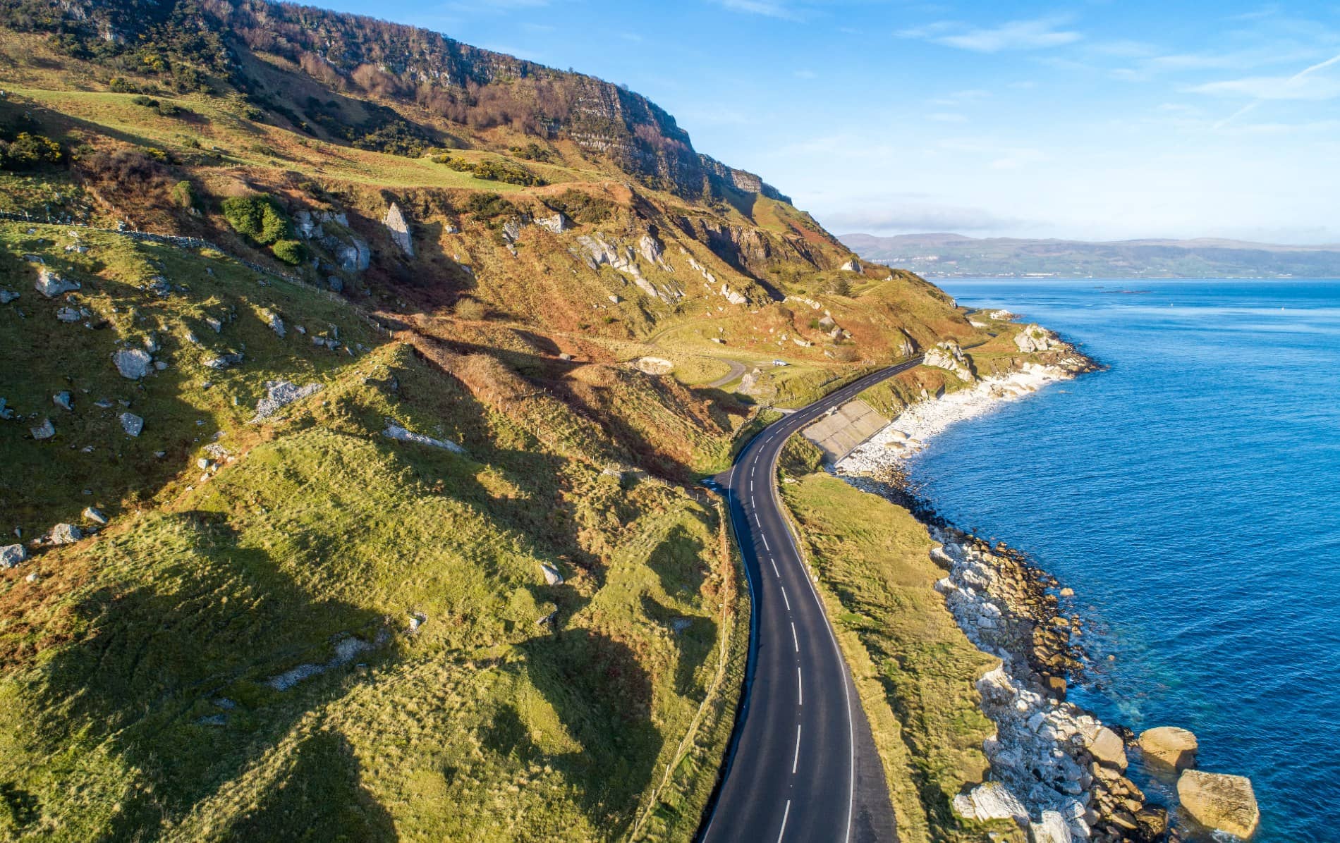 Coastal road on Causeway Coastal Route, Antrim, curving beside rocky cliffs and blue sea waters.