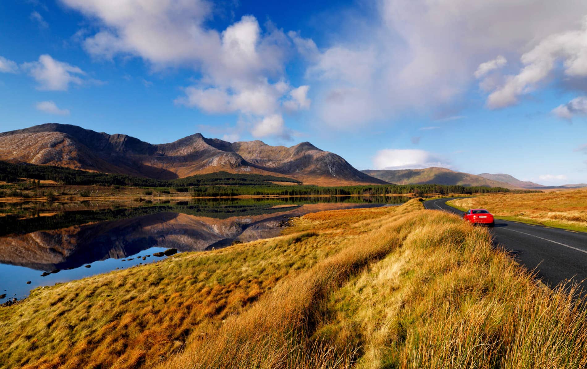 Red car driving past Twelve Bens mountains in Connemara, Galway, reflected in a calm lakeside view.