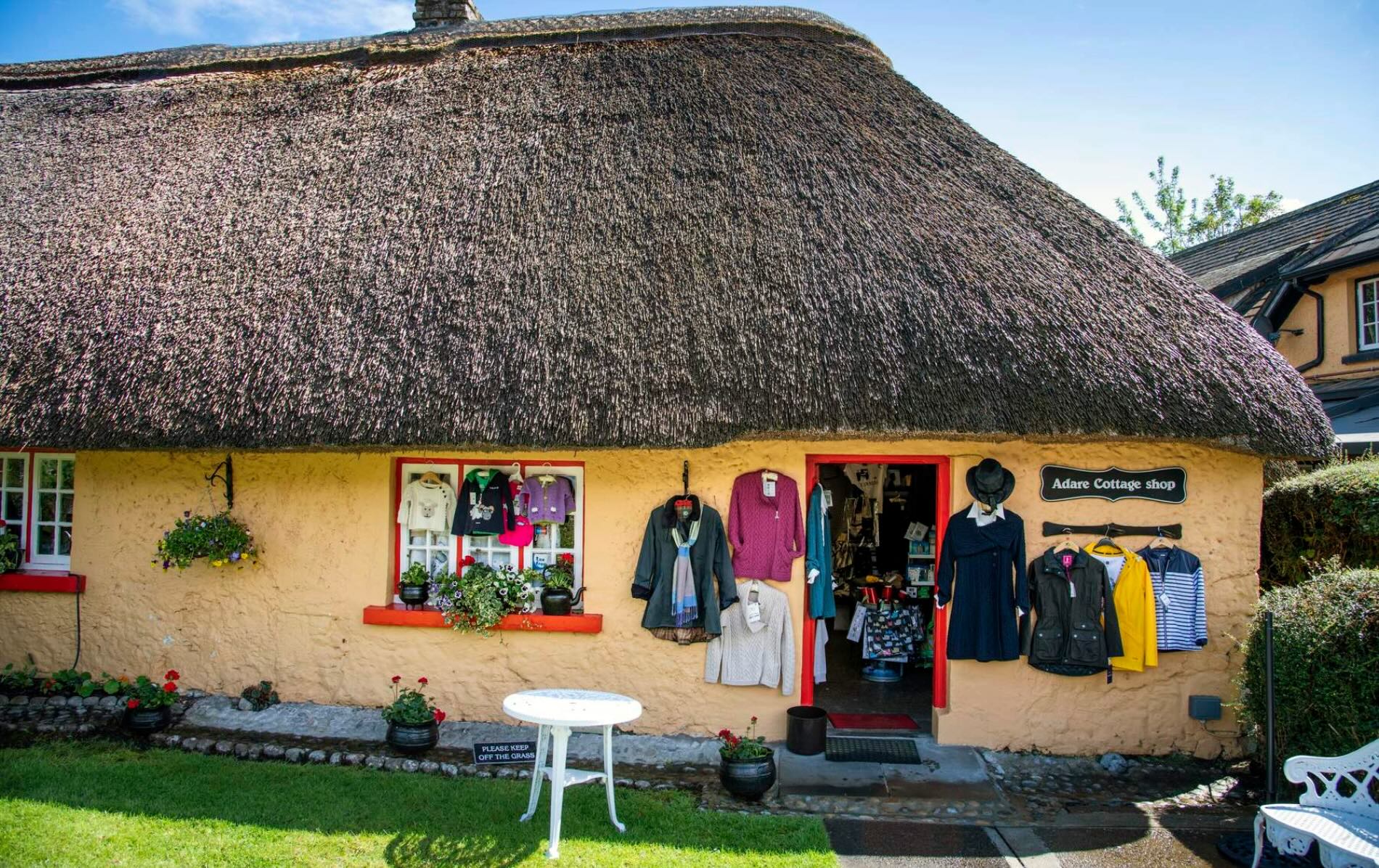 Thatched cottage front of Adare Cottage Shop with colourful knitwear, jackets, and hats displayed outside.