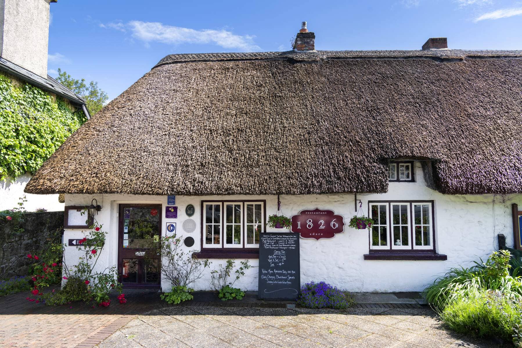 Thatched cottage housing the 1826 Adare Restaurant with white façade, flowers, and a chalkboard menu outside.