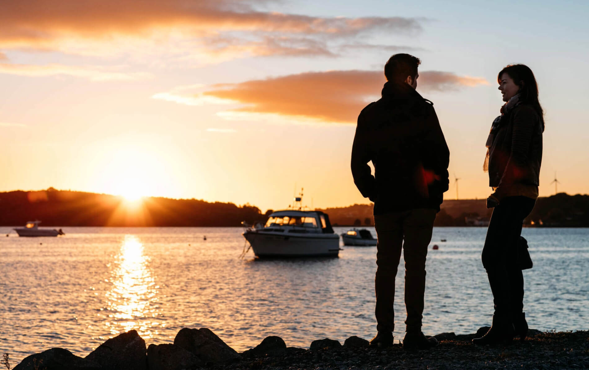 A couple stand by the water’s edge at sunset in Cobh, with boats moored in the harbour and golden light on the water.