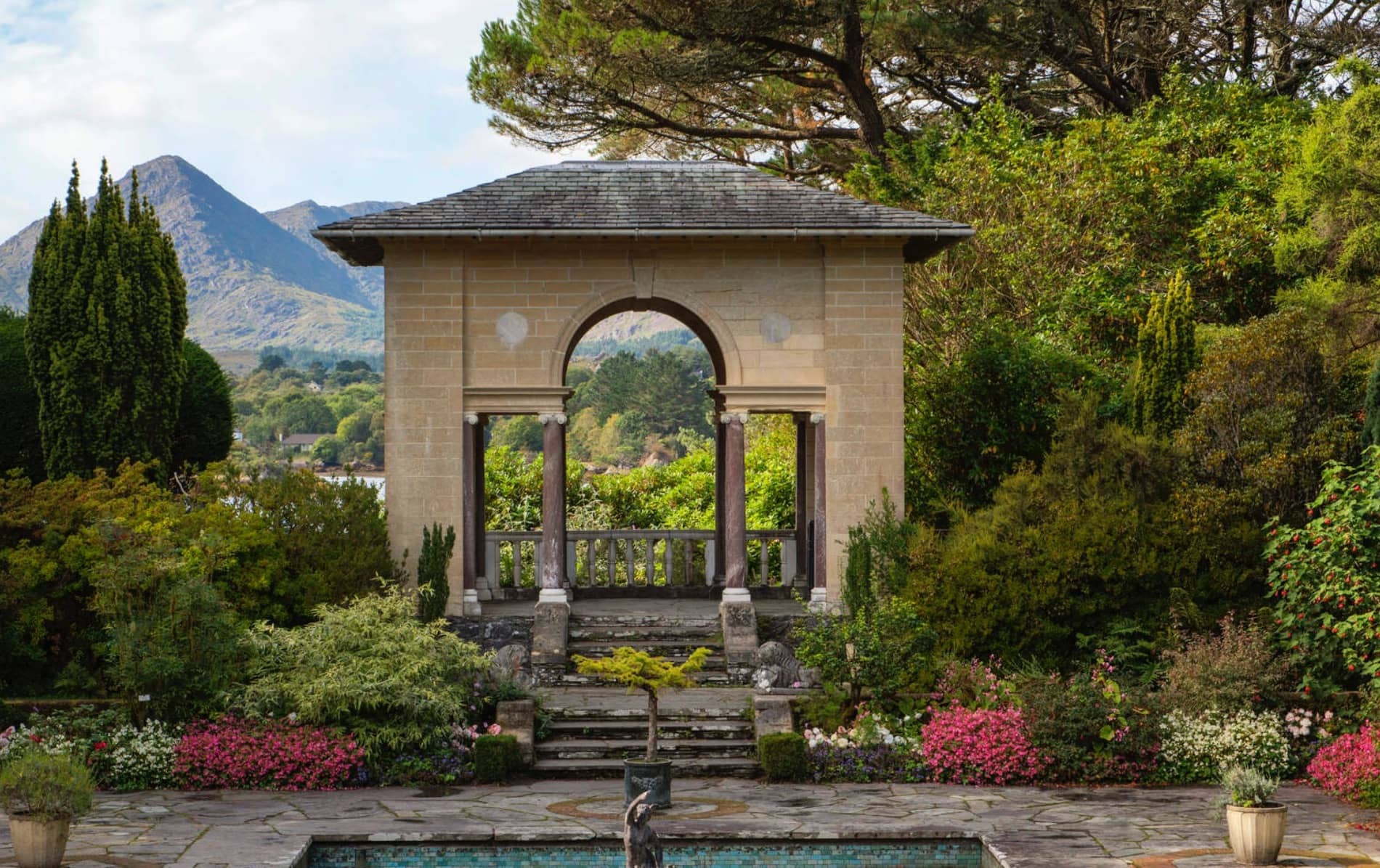 A formal garden on Garnish Island, Cork, featuring an elegant stone archway, manicured flowerbeds, and mountains in the background.