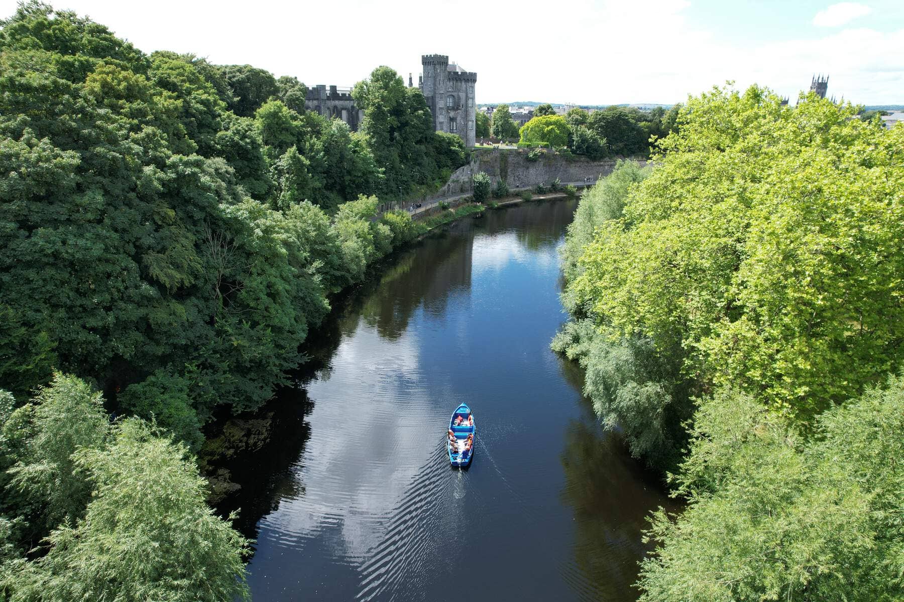 Small blue boat with passengers gliding along the River Nore, flanked by lush greenery and Kilkenny Castle.