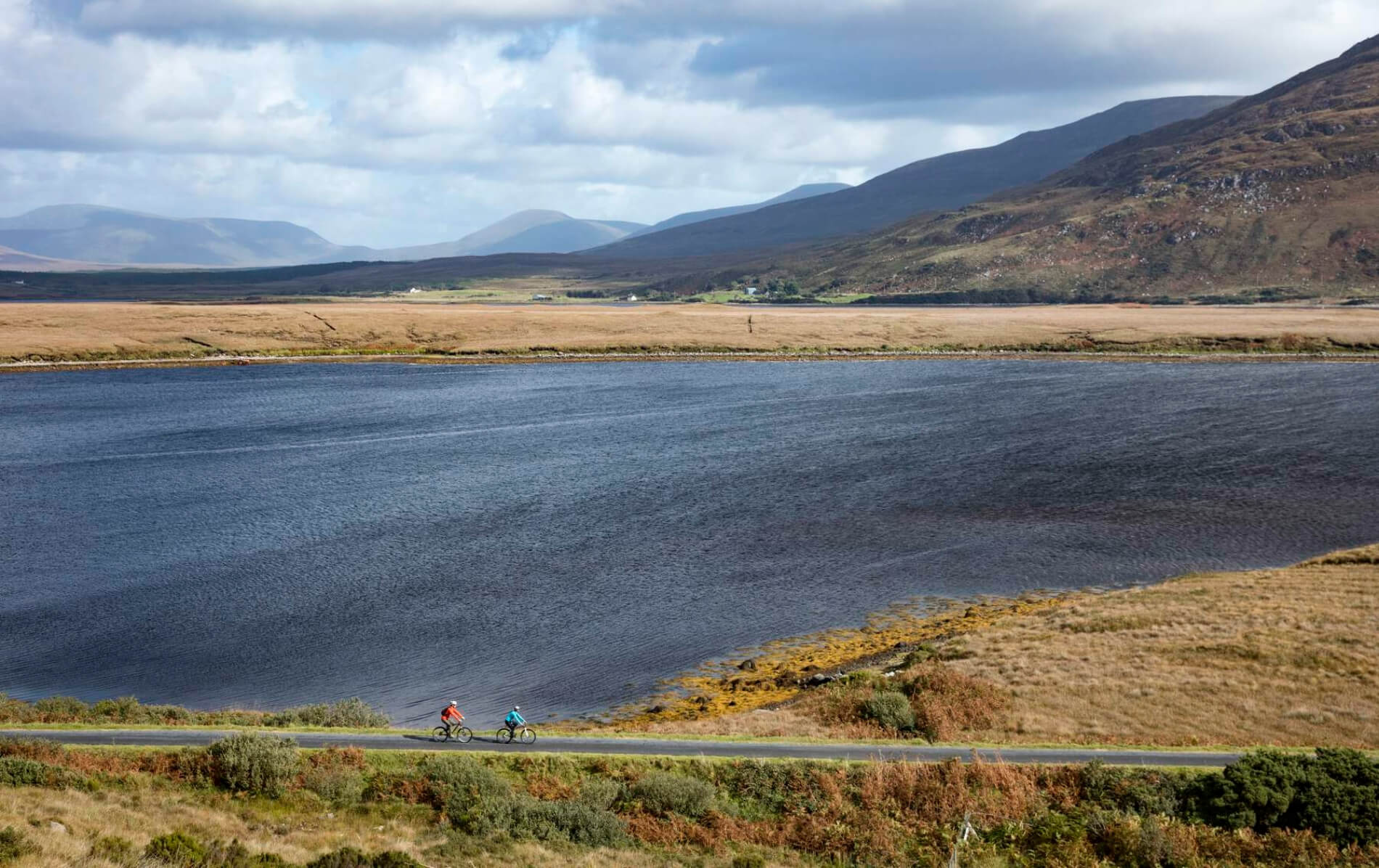Two cyclists ride a trail beside a vast lake, framed by open bogland and distant mountains.