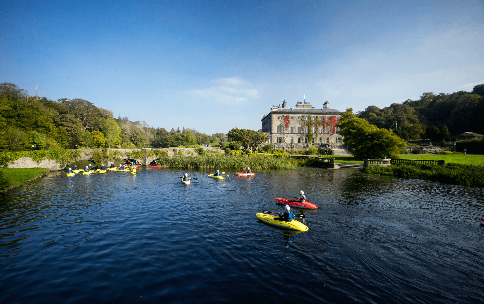 A group of kayakers paddle on calm river waters near a stately home surrounded by green parkland.