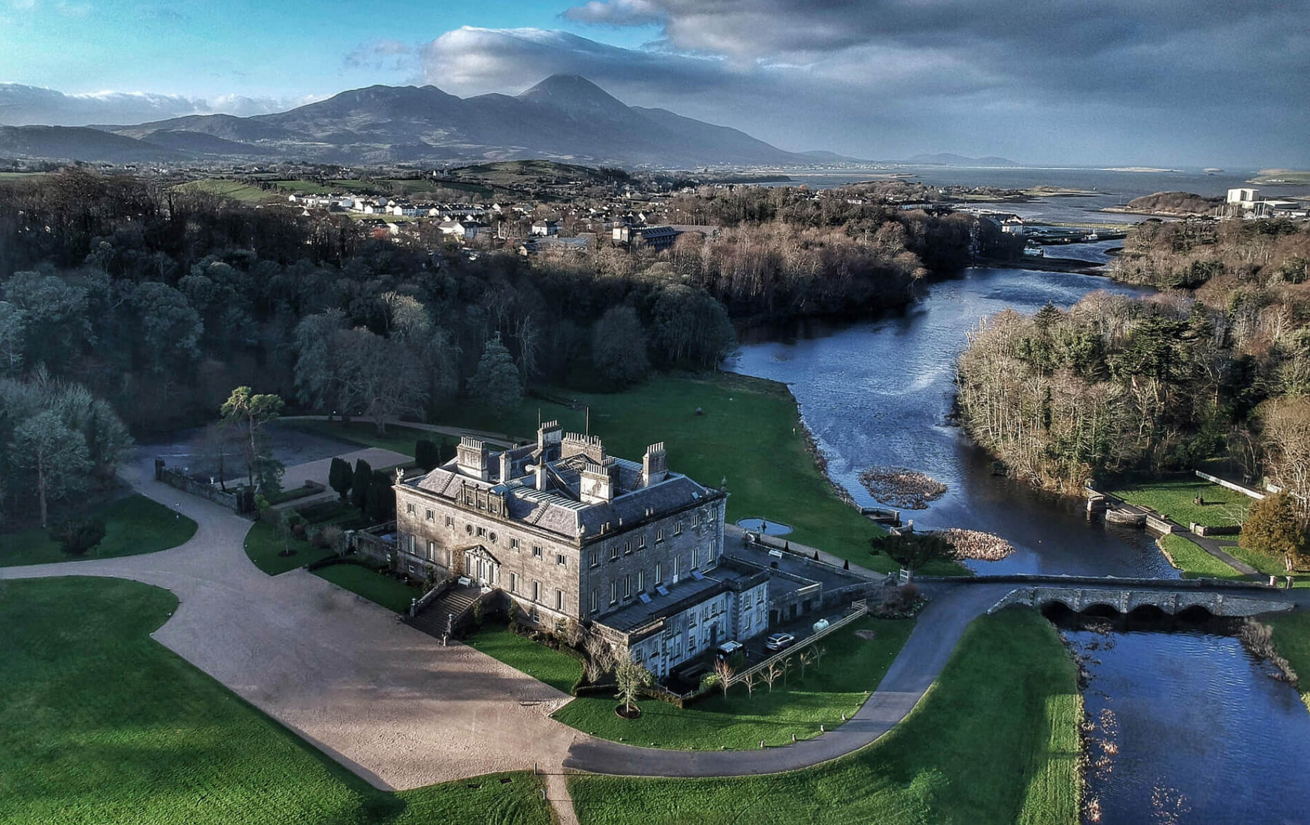 Aerial view of Westport House beside a river, with forest, village rooftops, and Croagh Patrick in the background.