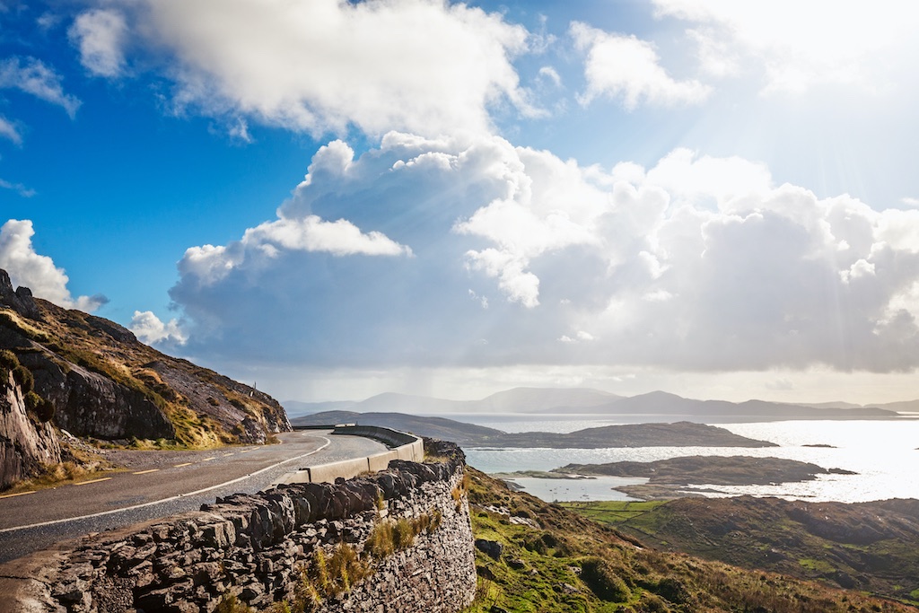 Scenic coastal road on the Ring of Kerry, curving along rocky cliffs with ocean views and dramatic cloud formations over County Kerry.