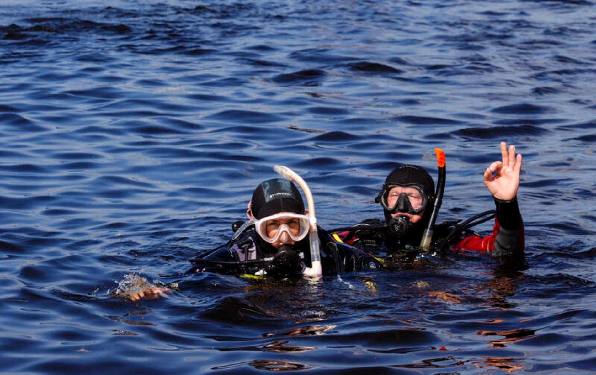 Two scuba divers in wetsuits and masks float in dark water at Dromineer, County Tipperary, one signalling OK with hand.