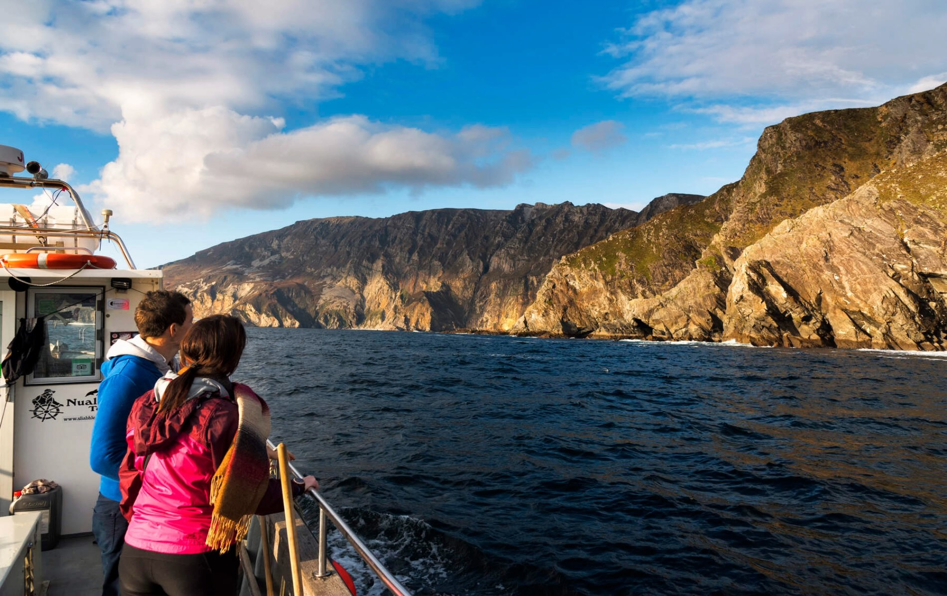 Couple on boat tour admiring Slieve League cliffs in County Donegal rising steeply above Atlantic Ocean.
