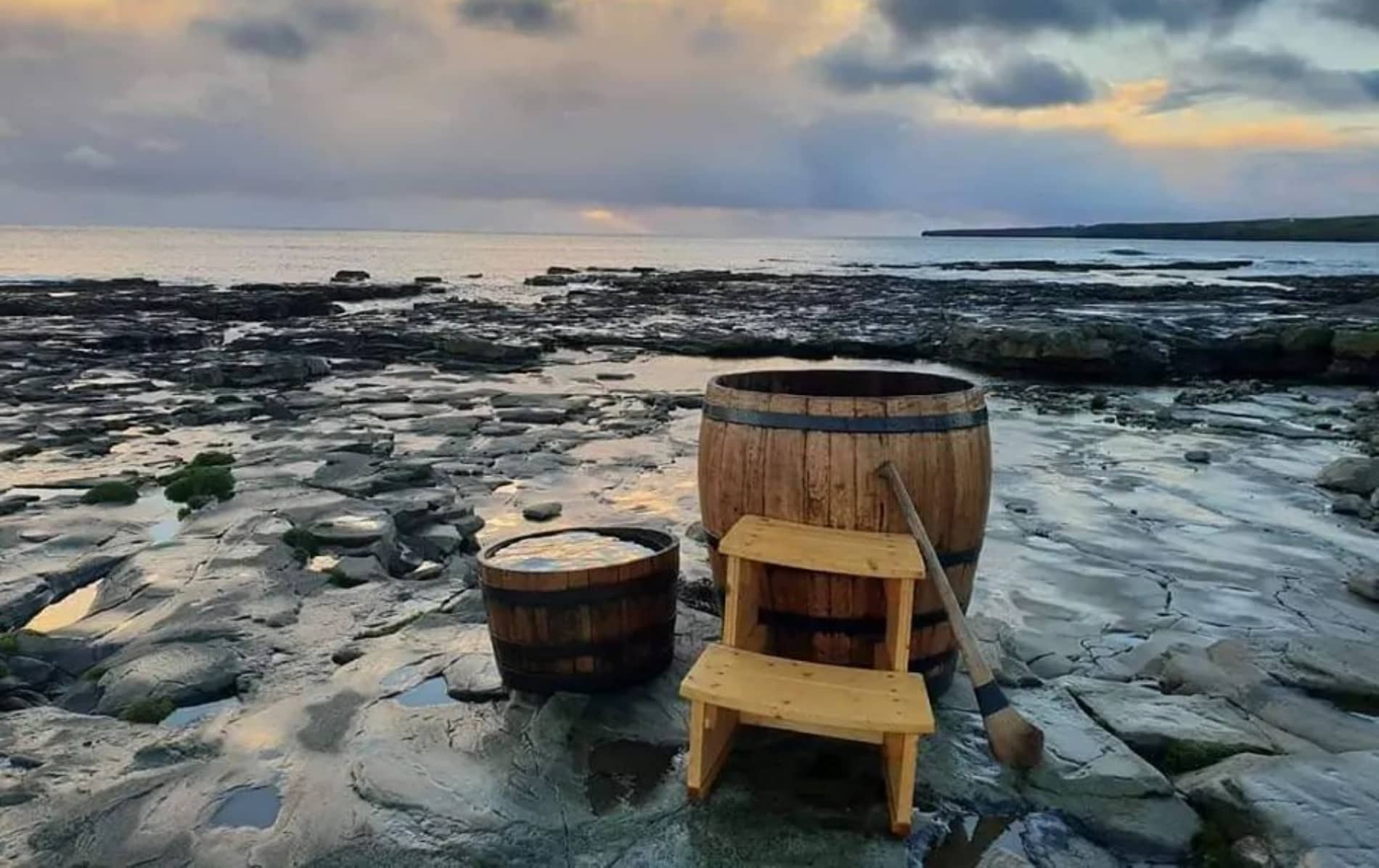 Two wooden barrel tubs with a paddle on rocky shoreline facing calm Atlantic waves at dusk.