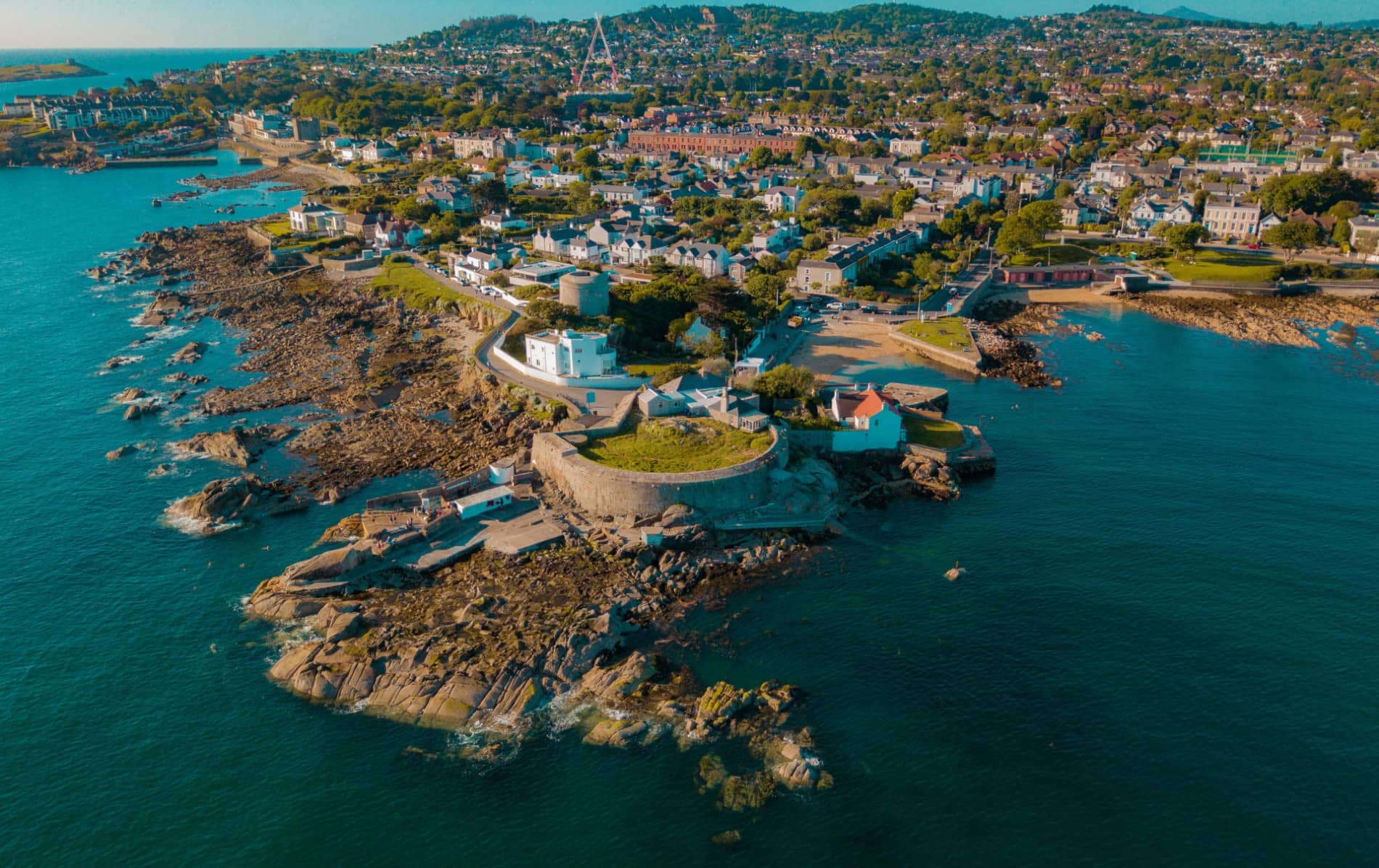 Aerial view of Dalkey coastline and Forty Foot bathing spot with swimmers on rocks in blue sea.