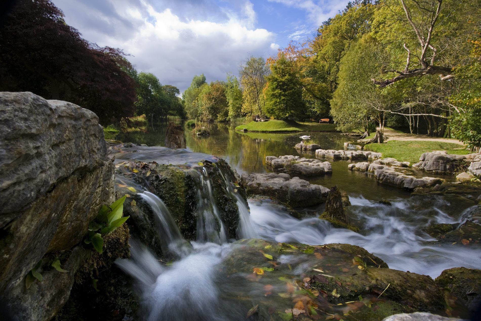 Small waterfall flows into a calm pond surrounded by trees and gardens in Ireland.