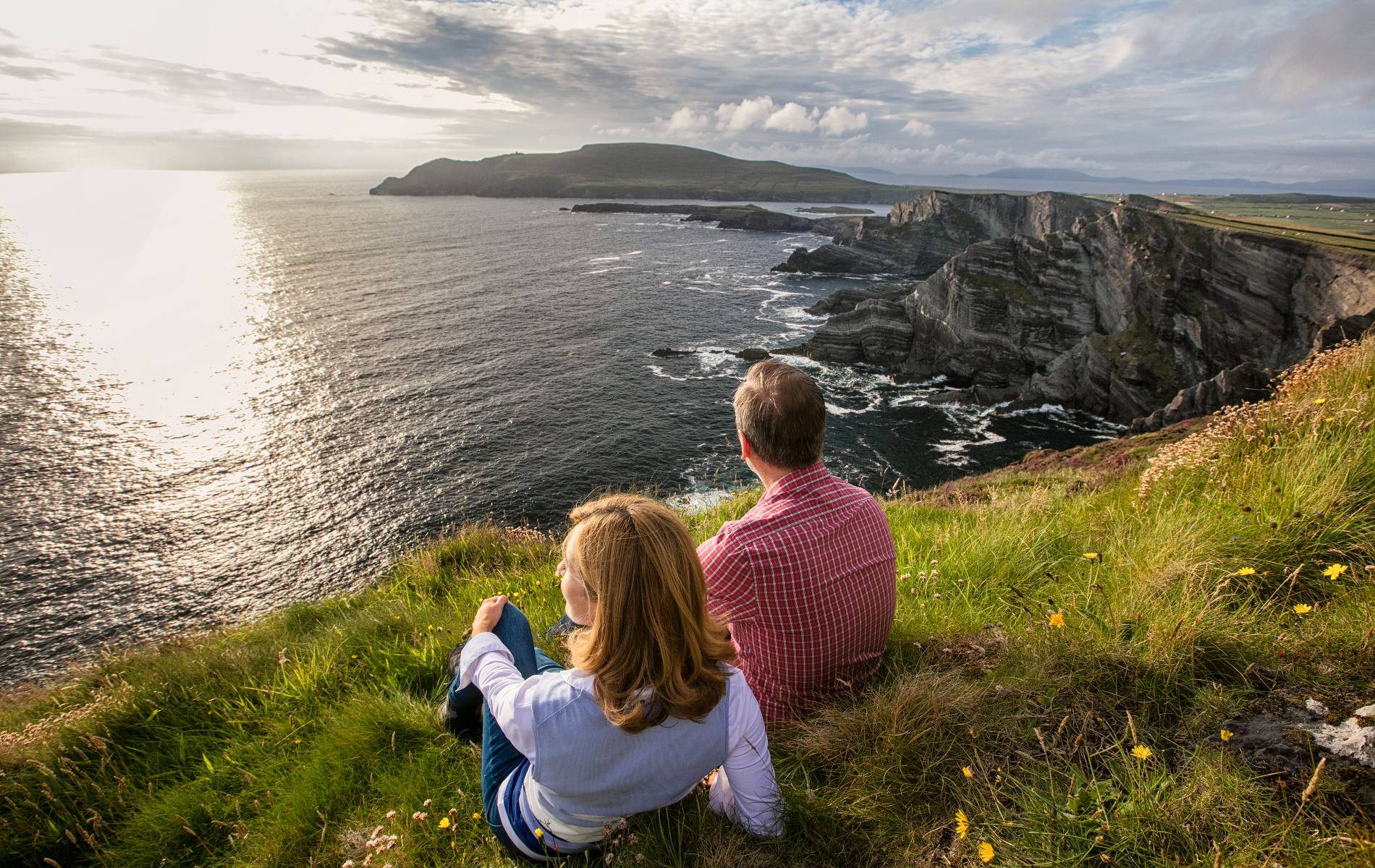 Couple sitting on a grassy cliff at sunset overlooking rugged cliffs and the Atlantic Ocean in County Kerry.