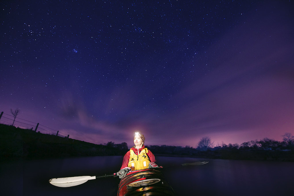 Kayaker with headlamp paddling on the Roe River beneath a vivid night sky of stars.