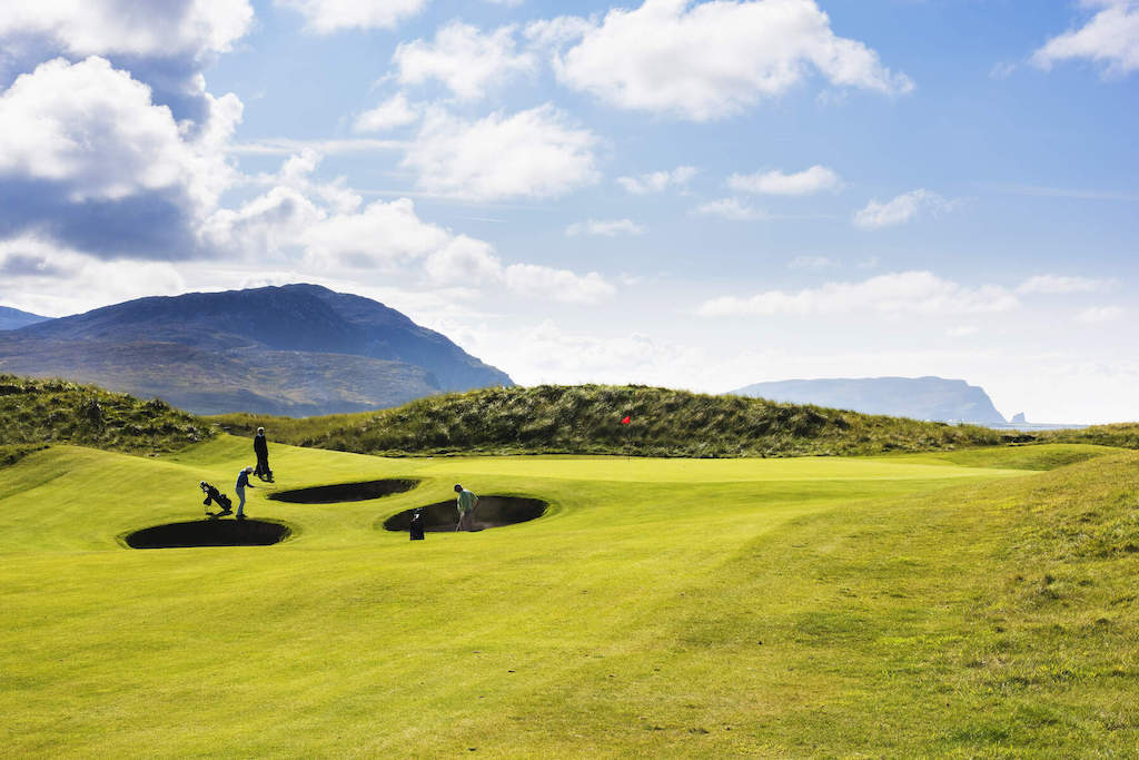 Golfers play near deep bunkers on a green with mountain and coastal views at Ballyliffin Golf Course, Donegal.