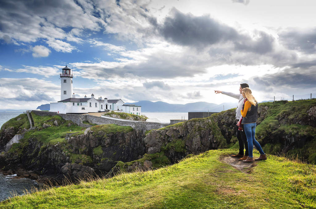 Two people stand on a grassy cliff pointing toward Fanad Lighthouse on Donegal’s dramatic Atlantic coastline.
