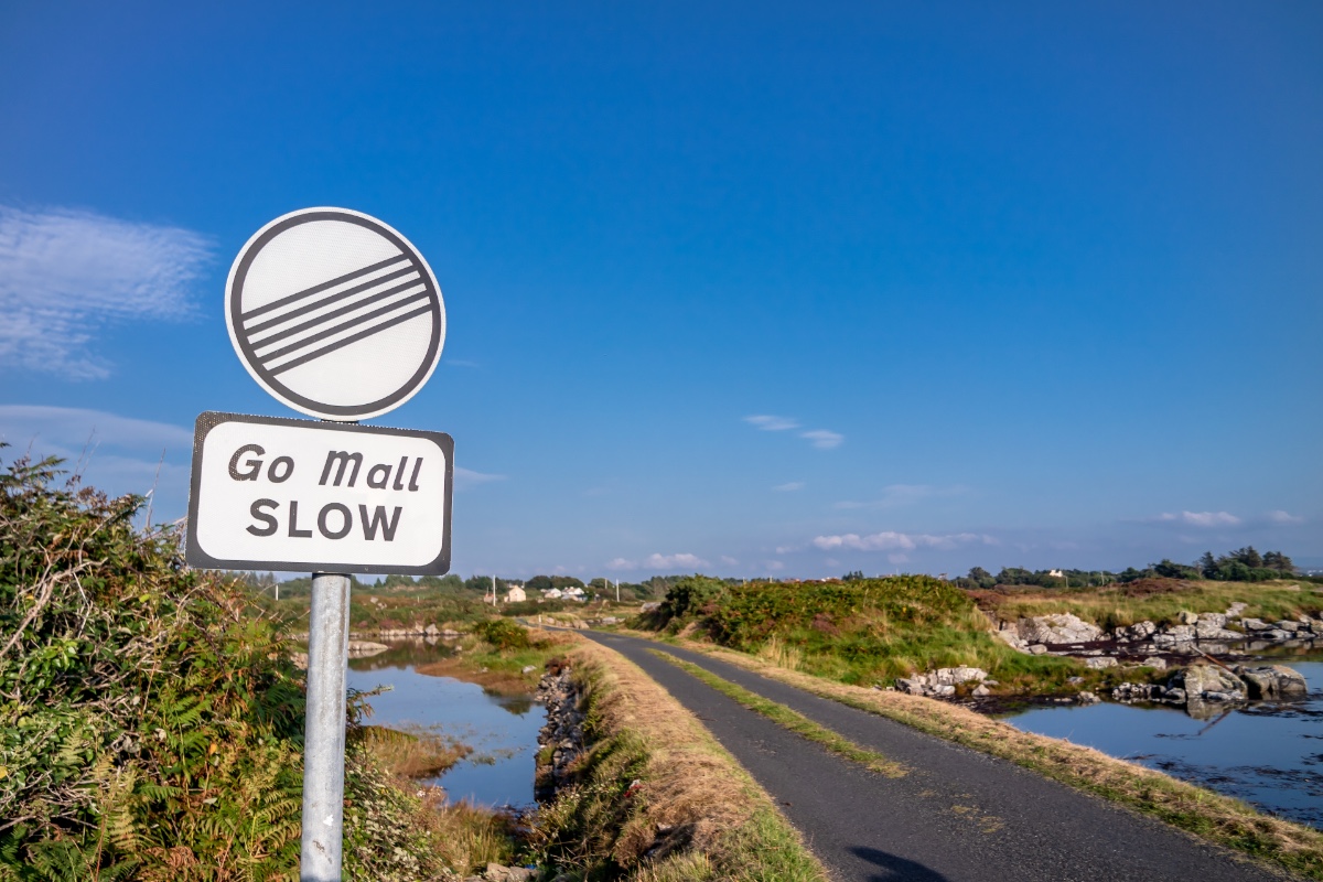 Bilingual Irish road sign reading ‘Go Mall Slow’ stands beside a rural road and waterway in County Kerry.