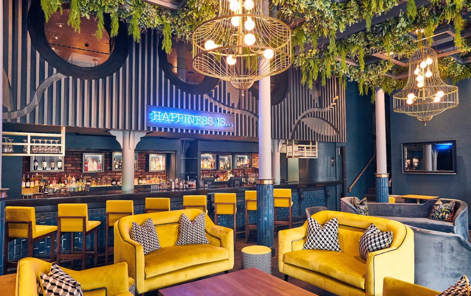 Lush cocktail bar with yellow armchairs, hanging plants and a neon ‘Happiness Is…’ sign in Malmaison, Belfast.