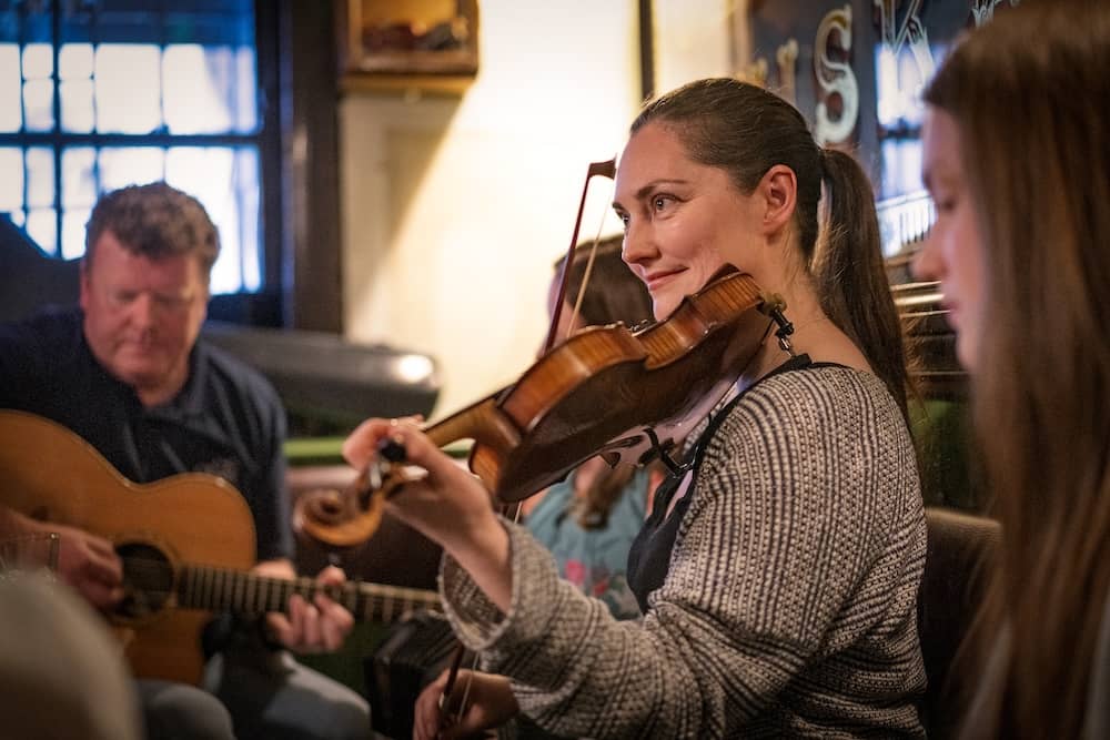 Musicians playing fiddle and guitar during a lively traditional music session in a Belfast pub.