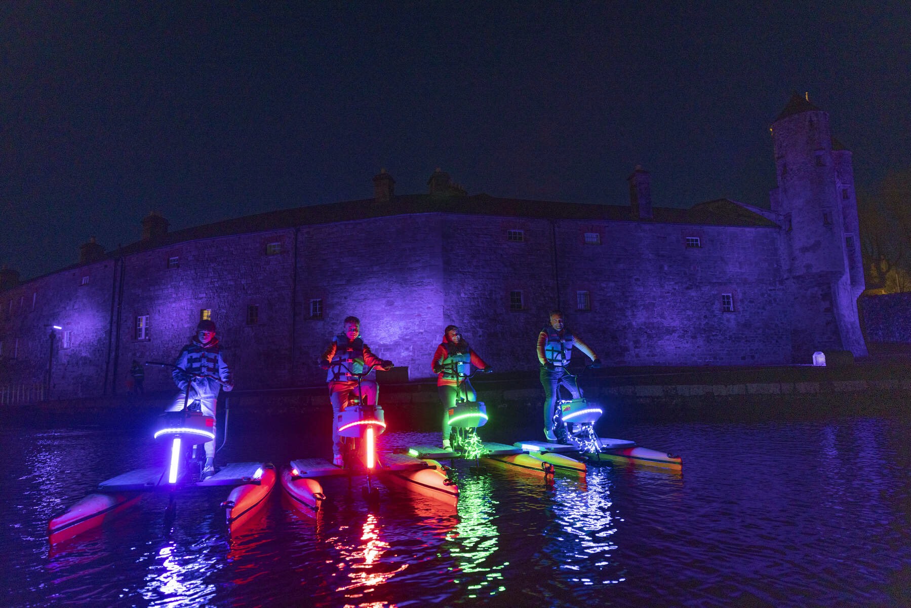 Group on illuminated water bikes with glowing neon lights in front of Enniskillen Castle lit purple at night.