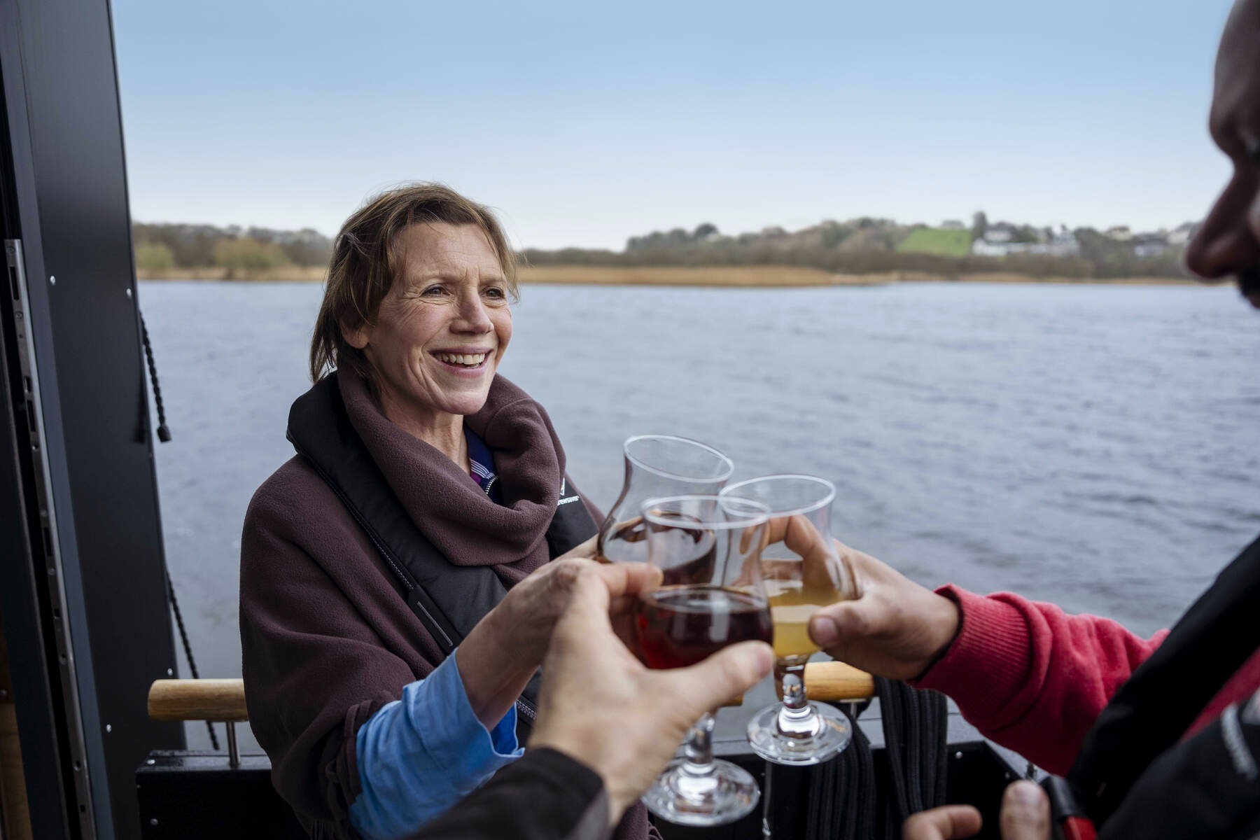 Woman smiling on boat as glasses of wine and juice are raised in a toast on Lough Erne, with shoreline in the distance.