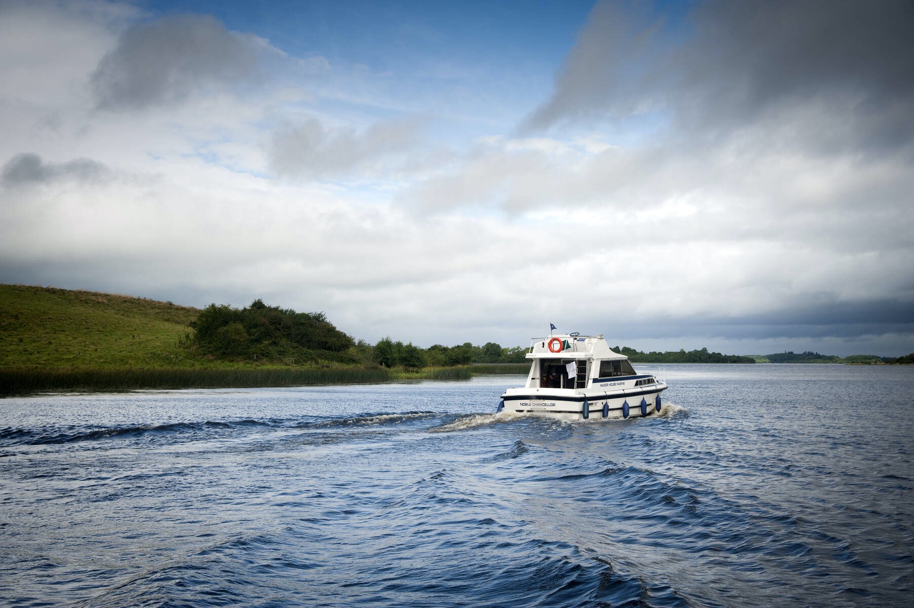 Small white boat sailing across the waters of Lough Erne surrounded by grassy hills and cloudy skies in County Fermanagh.
