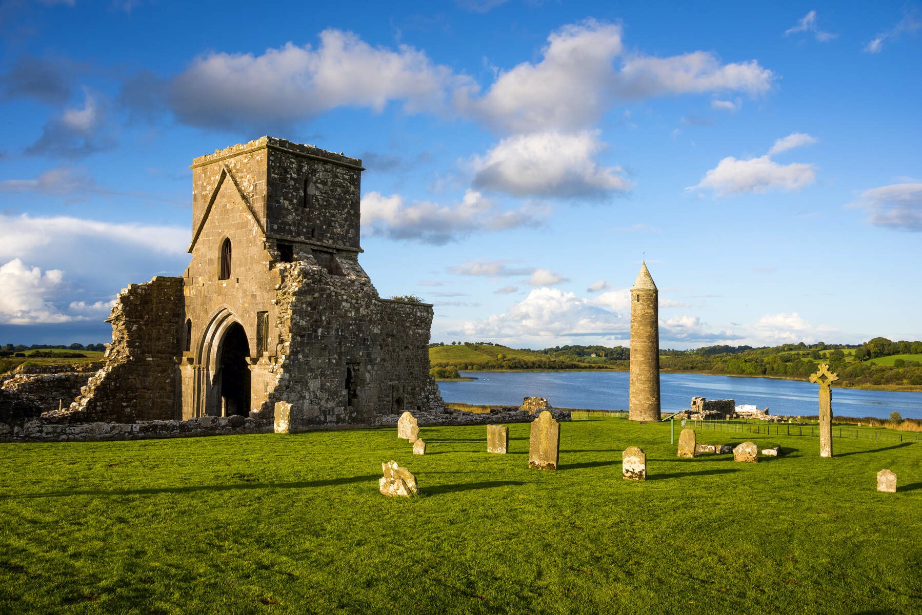 Stone ruins, round tower, and old gravestones at Devenish Monastic Site on Lough Erne with blue sky and water backdrop.