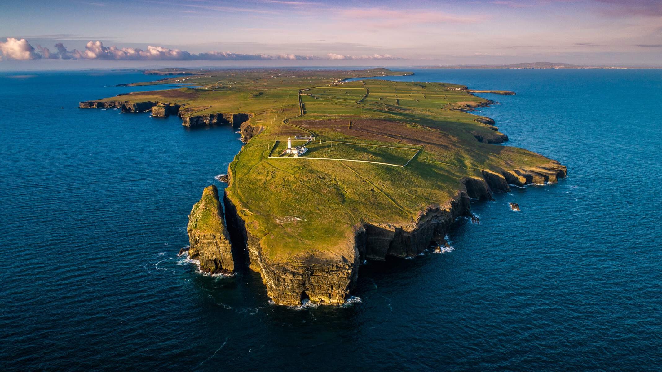 Aerial view of Loop Head Peninsula, County Clare, with cliffs, fields, and lighthouse surrounded by blue sea.
