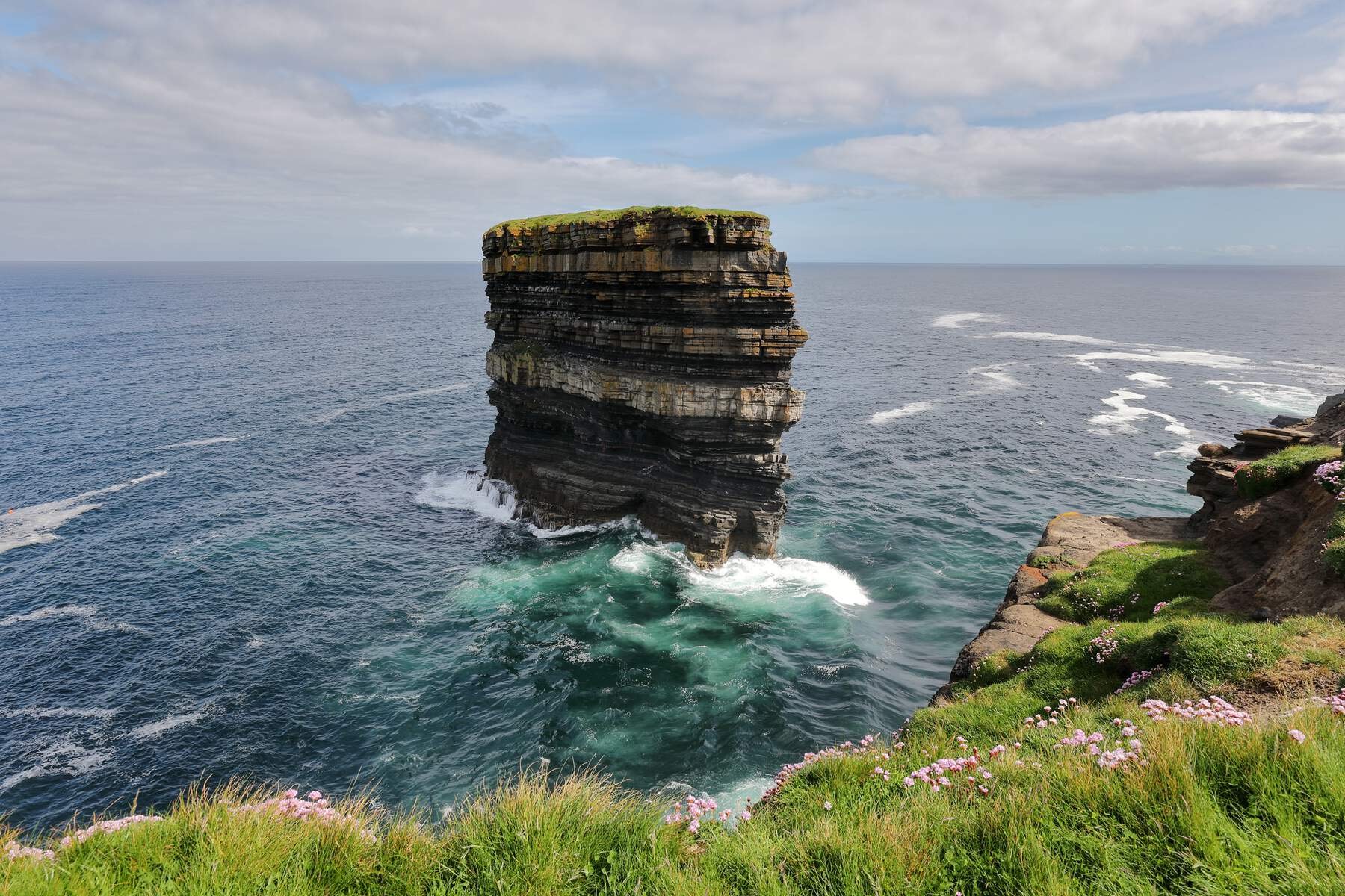 Vertical sea stack at Downpatrick Head in Mayo surrounded by turquoise waves and cliffs topped with grass.