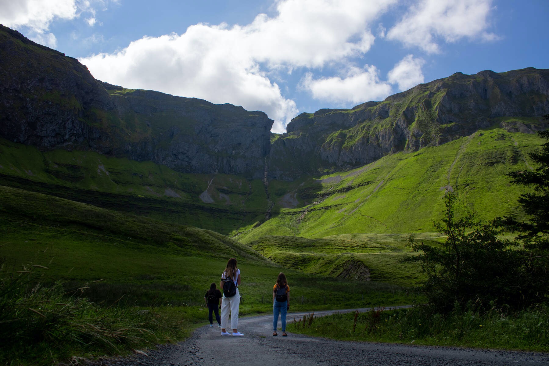 Three walkers on a path facing tall green cliffs under a blue sky in the remote Gleniff Horseshoe, County Sligo.