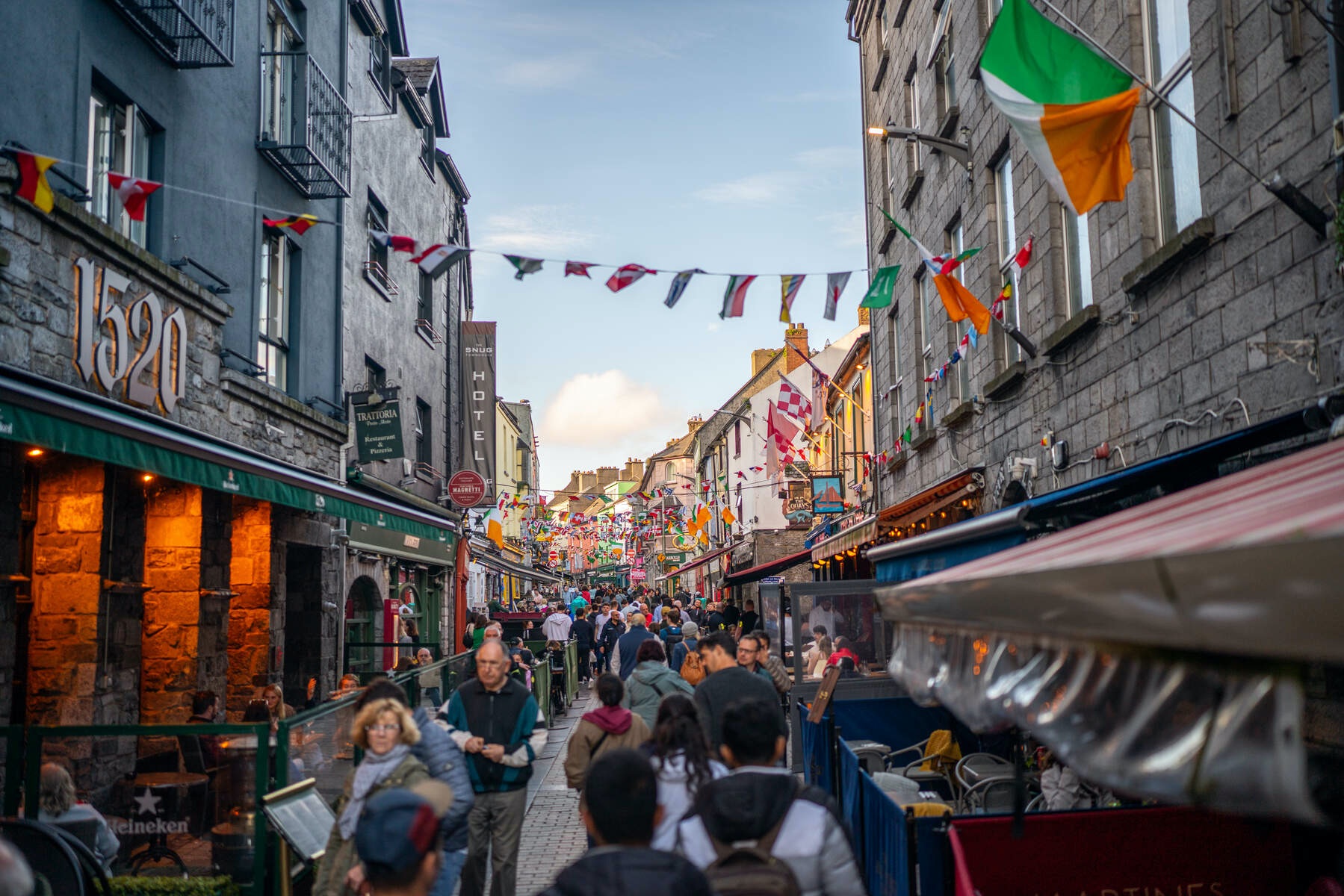 Colourful flags strung above a bustling pedestrian street in Galway city that is filled with people.