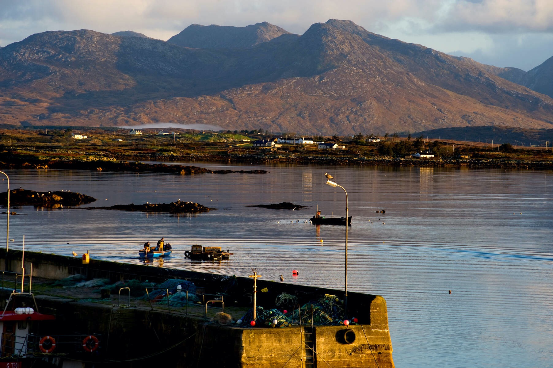 Quiet fishing pier at Roundstone, Galway, with boats and nets, set against calm waters and rugged mountain backdrop.
