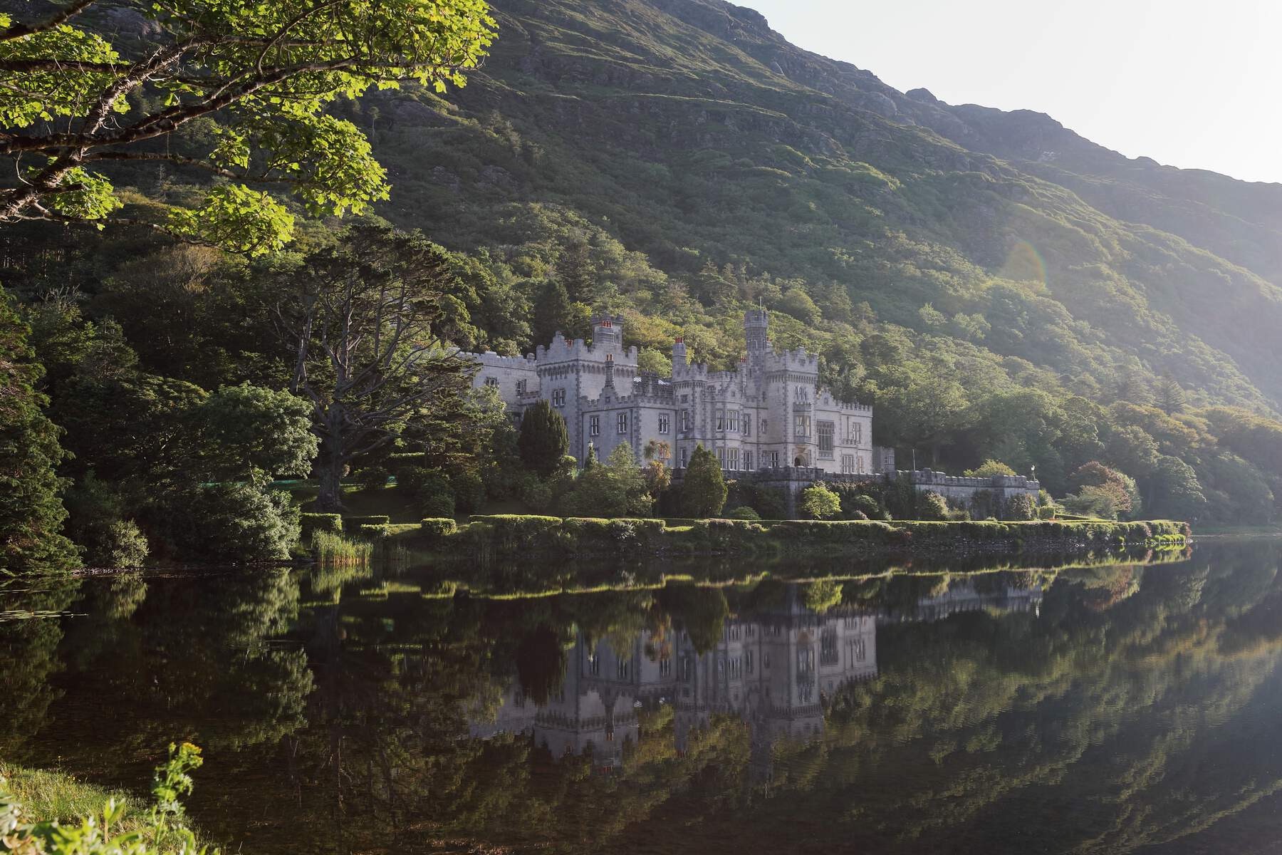 Kylemore Abbey in County Galway reflected in still water, surrounded by trees and green hillside.