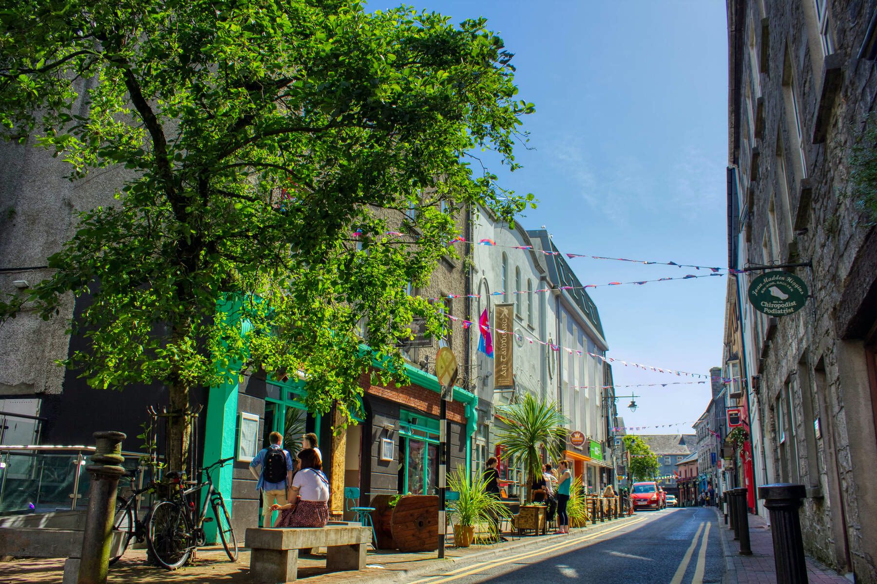Quiet street in Galway city with bunting, a tree in full leaf, and people outside shopfronts.