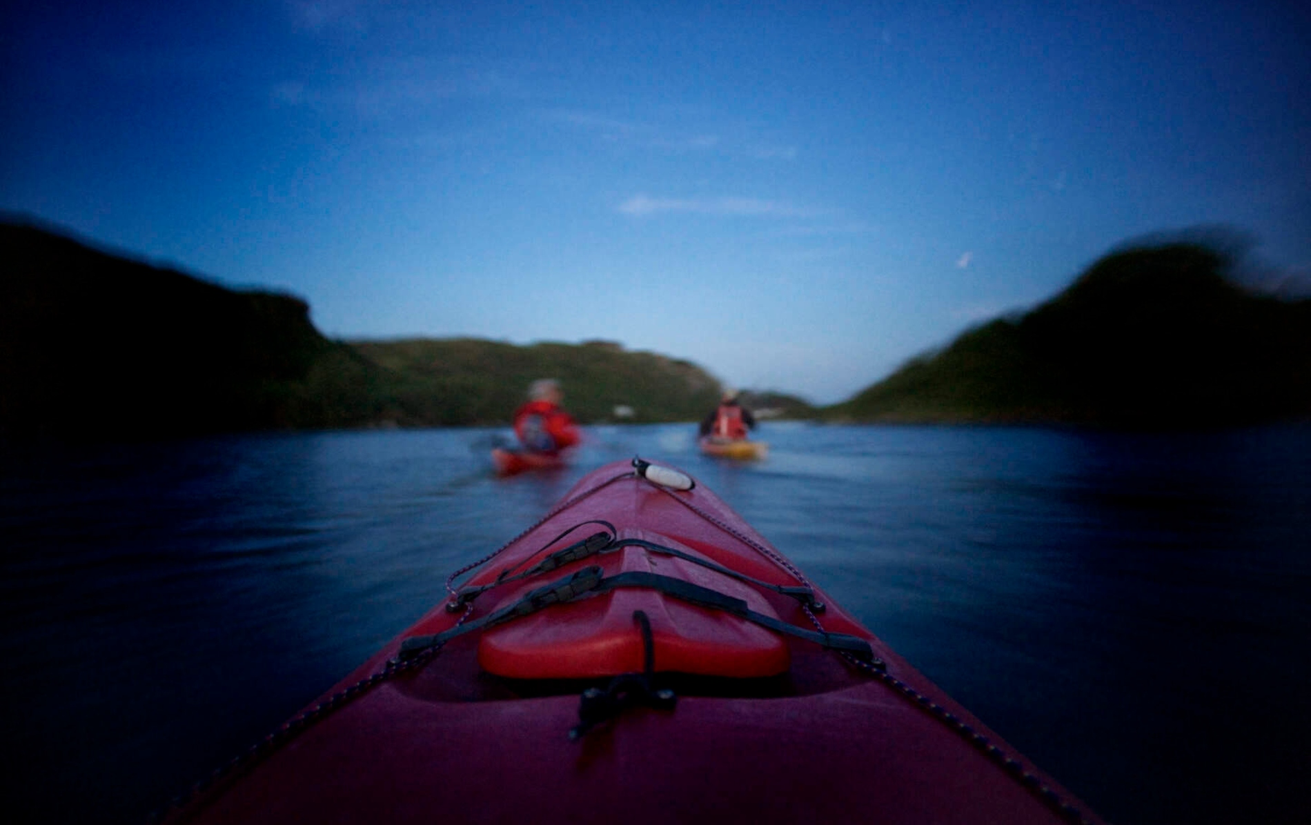 View from a red kayak at twilight on Lough Hyne in County Cork, with other kayakers paddling ahead between forested hills.