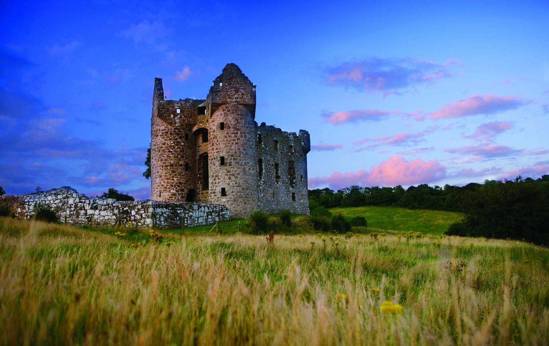 Ruins of Monea Castle in County Fermanagh, standing tall on a grassy field at dusk with a pink-tinged sky in the background.