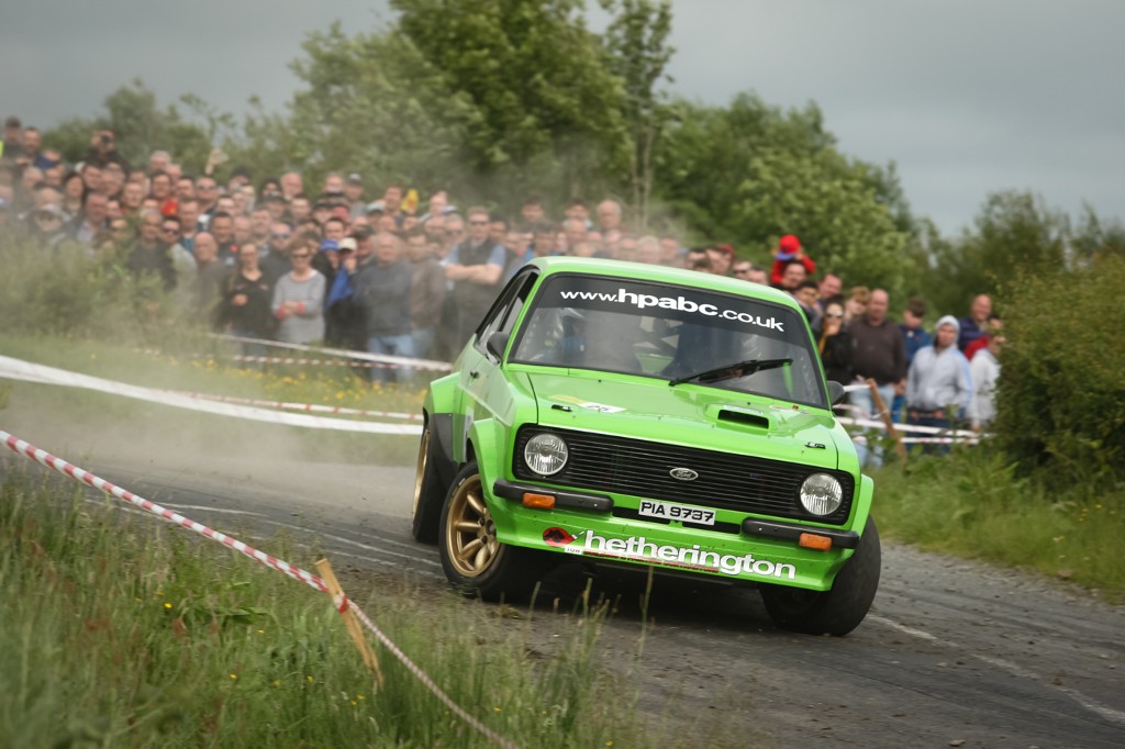 A green Ford rally car drifts through a turn on a wet country road, kicking up spray in front of a crowd.