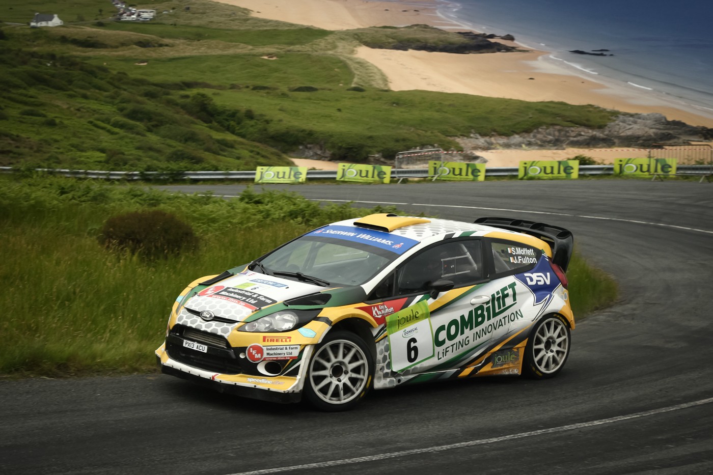 A rally car turns a coastal road corner at speed with a beach and sea visible in the background.